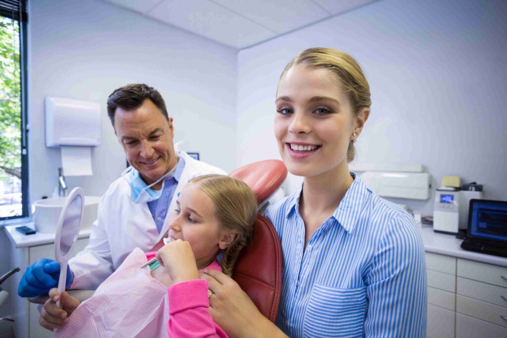 Dentist examining a child’s teeth while her mother smiles beside her.